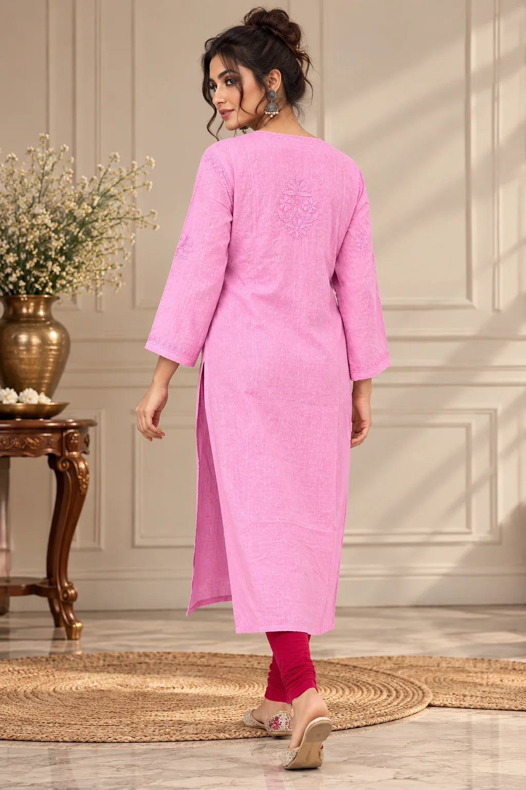 A woman wearing a pink three-quarter sleeve kurta with red leggings and beige flat shoes, shown from the back in an indoor setting with a decorative table and vase.