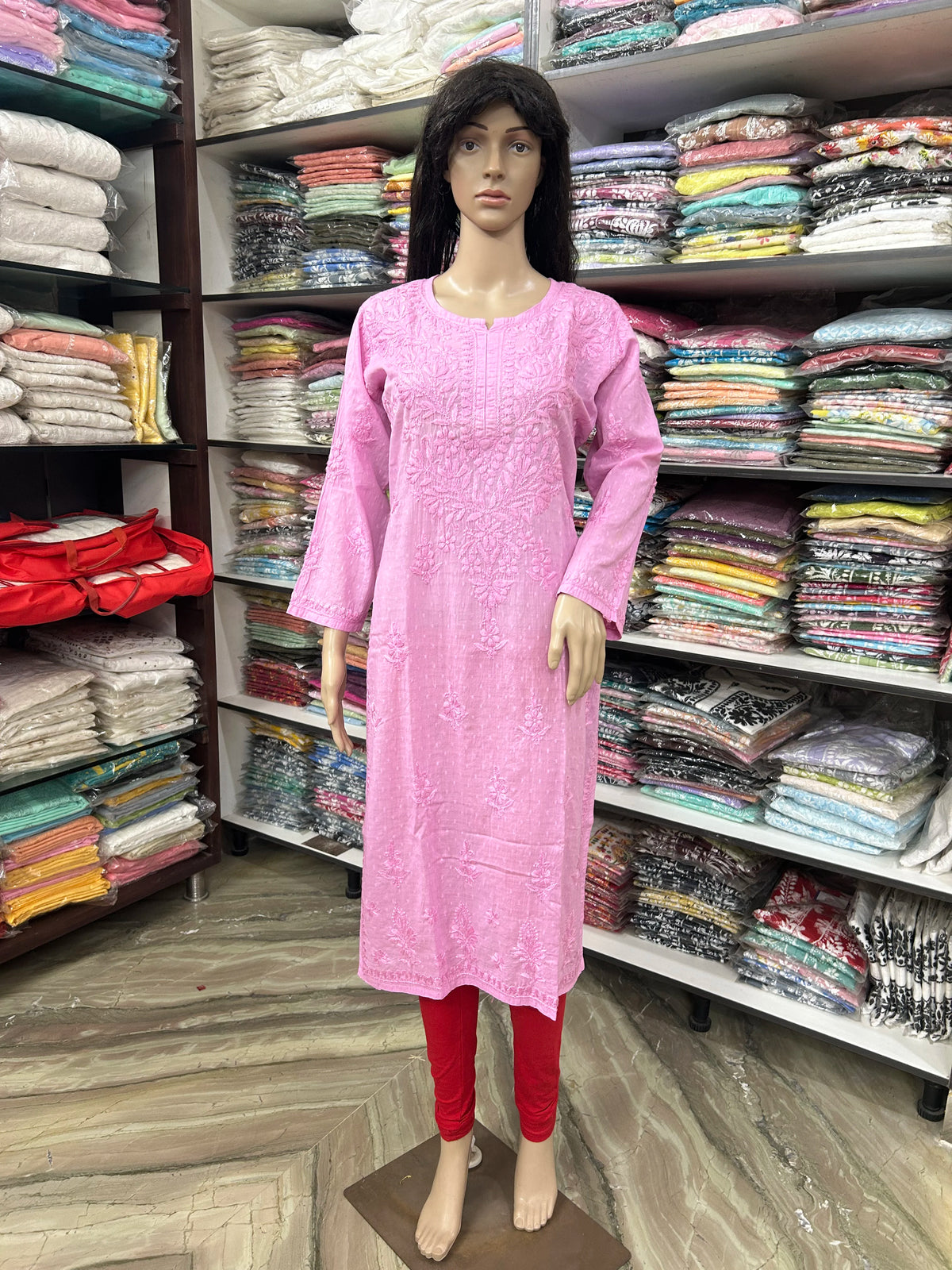 A pink embroidered long kurta displayed on a female mannequin, paired with red leggings, set against a background of shelves filled with folded clothing.