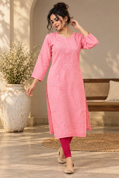A woman models a pink embroidered kurta with three-quarter sleeves and side slits, paired with magenta leggings and gold embellished flat shoes in a warm, sunlit indoor setting.