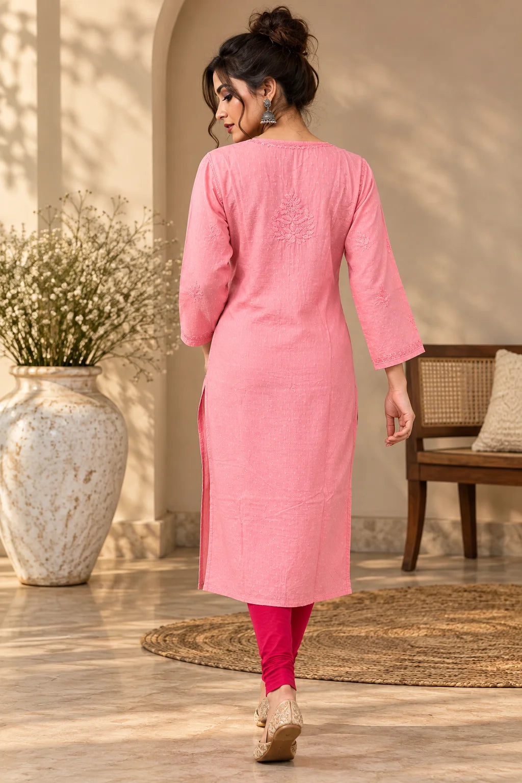 A woman wearing a pink three-quarter sleeve kurta with side slits, paired with bright pink leggings and beige wedge sandals, shown from behind in a warm, sunlit indoor setting with a large vase and woven rug.