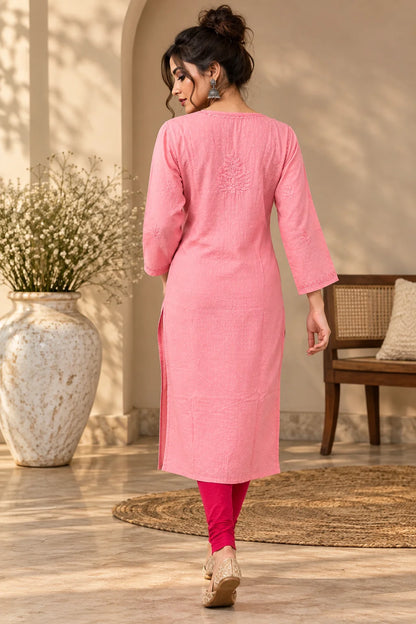 A woman wearing a pink three-quarter sleeve kurta with side slits, paired with bright pink leggings and beige wedge sandals, shown from behind in a warm, sunlit indoor setting with a large vase and woven rug.