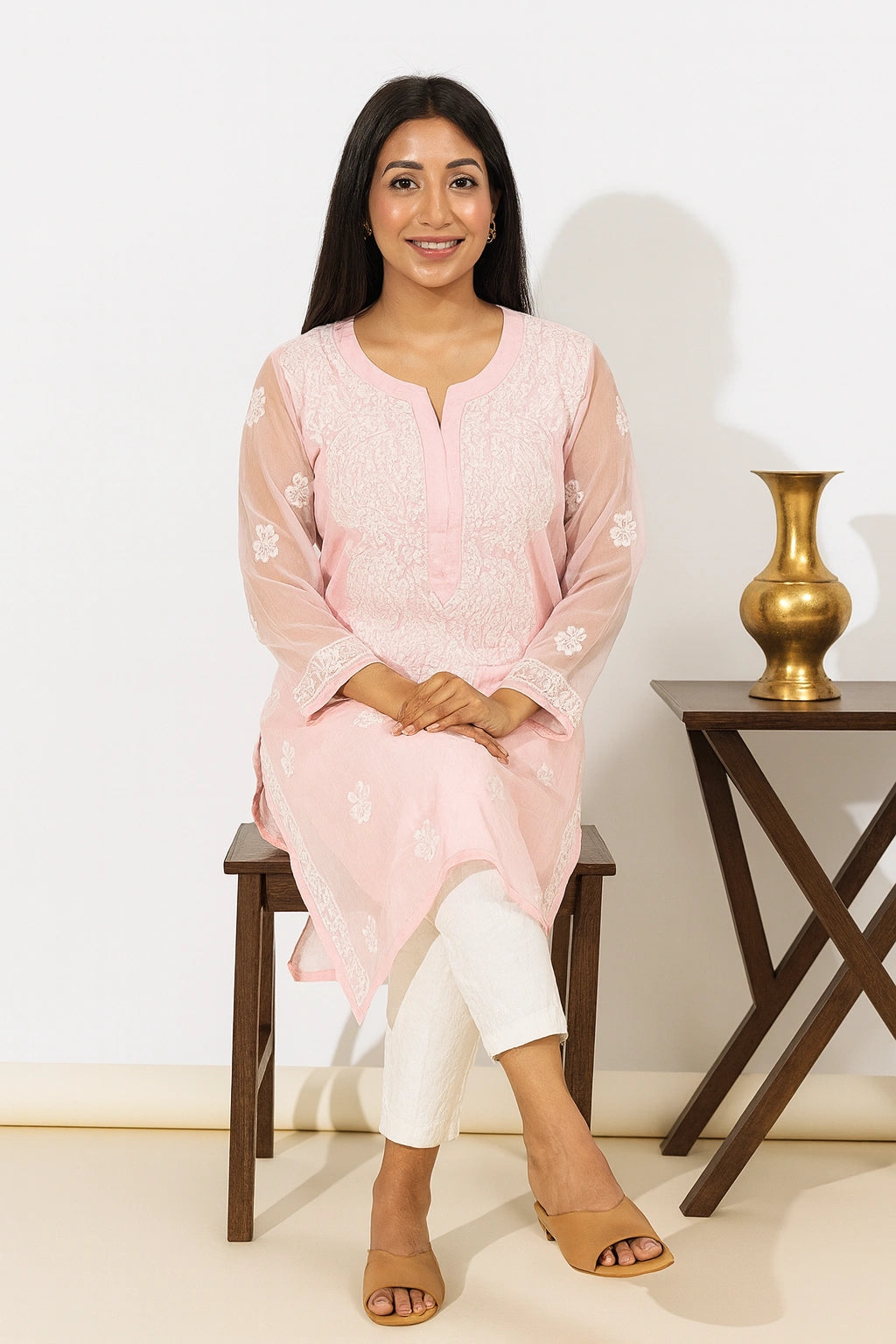 A woman sits on a dark wooden stool wearing a light pink embroidered kurta with white pants and tan sandals. The background is a clean white wall with a small wooden side table holding a gold vase.
