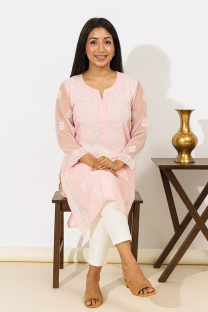 A woman sits on a dark wooden stool wearing a light pink embroidered kurta with white pants and tan sandals. The background is a clean white wall with a small wooden side table holding a gold vase.