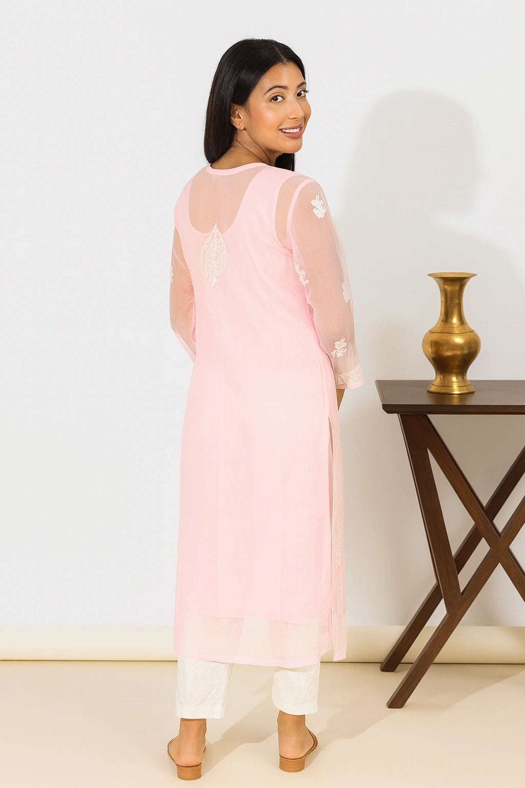 Lucknowi chikankari A woman modeling a light pink kurta with white pants in a bright, modern studio setting with decorative white pedestals.