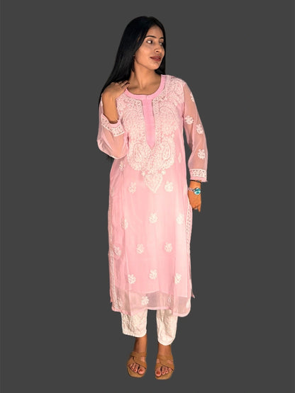 A woman modeling a light pink embroidered kurta with white floral patterns and matching white pants, standing against a solid dark gray background.