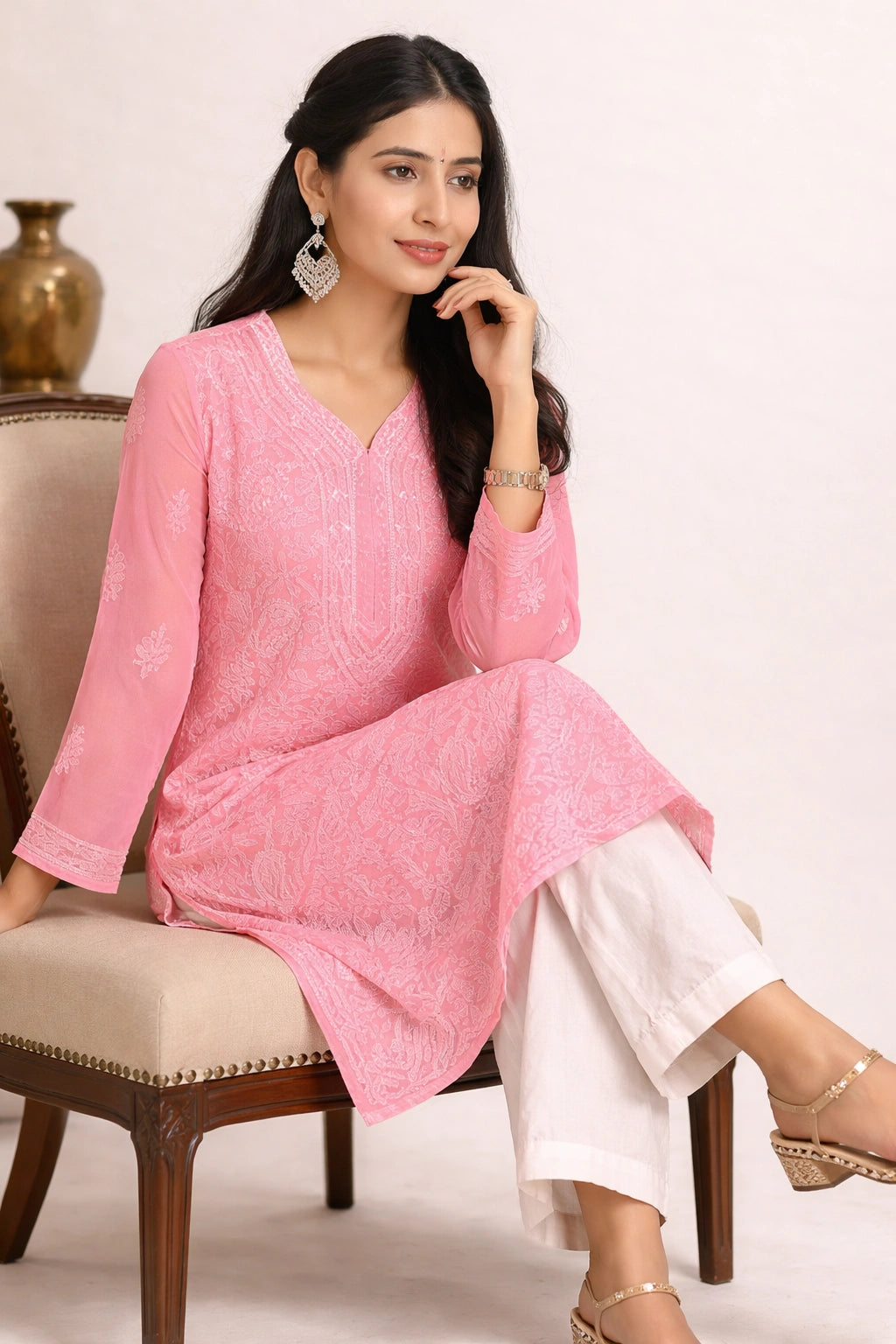 A woman seated on a beige upholstered chair wearing a pink embroidered kurta with white pants, against a plain light background.