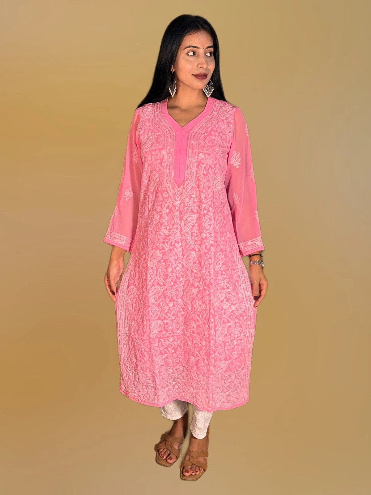 A woman modeling a light pink embroidered kurta with white floral patterns and matching white pants, standing against a solid dark golden background.