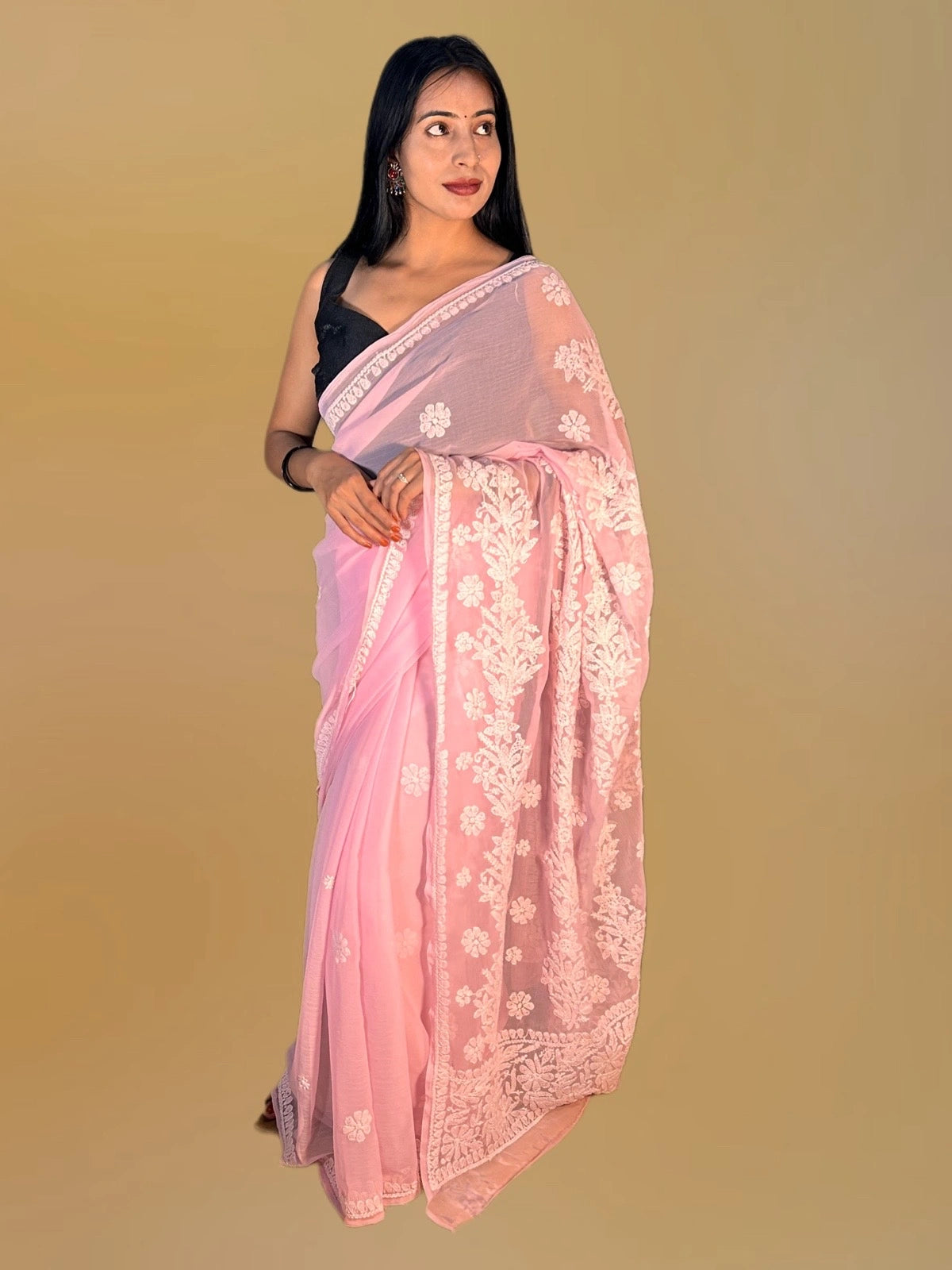 A woman modeling a light pink saree with white floral embroidery and a black sleeveless blouse, set against a solid beige background.