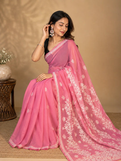A woman models a vibrant pink saree with intricate white floral embroidery along the border and pallu, paired with a black blouse, set against a warm beige background.