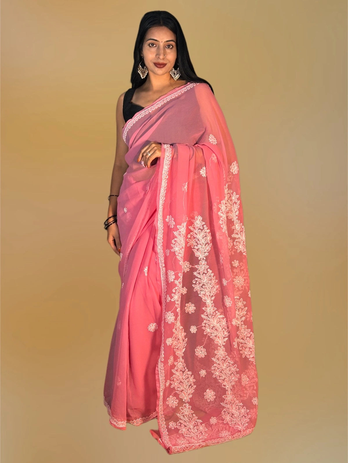 A woman modeling a pink saree with white floral embroidery and a solid pink border, paired with a black sleeveless blouse, against a solid beige background.