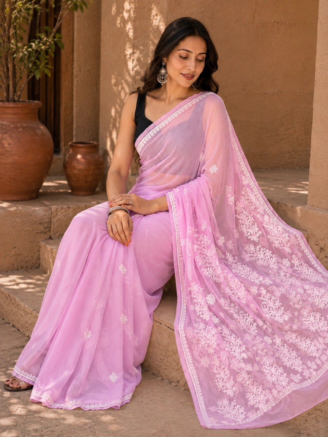 A woman wearing a light pink embroidered saree with a black sleeveless blouse, sitting on stone steps in front of a tan wall with terracotta pots.