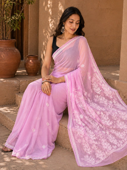 A woman wearing a light pink embroidered saree with a black sleeveless blouse, sitting on stone steps in front of a tan wall with terracotta pots.