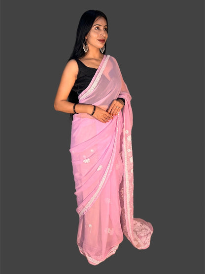 A woman modeling a light pink saree with white floral embroidery and a white border, paired with a black sleeveless blouse, against a solid dark gray background.