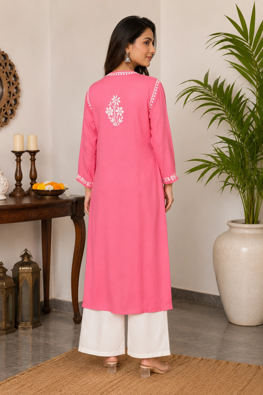 Woman wearing a pink kurta with white embroidery in a room with a table and plant.