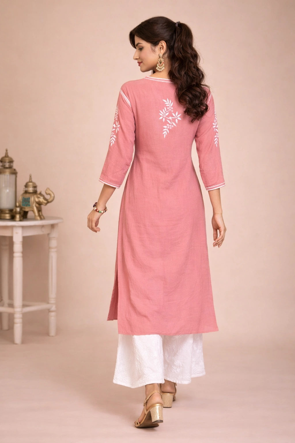 Woman wearing a pink kurta with white embroidery in a decorated room.