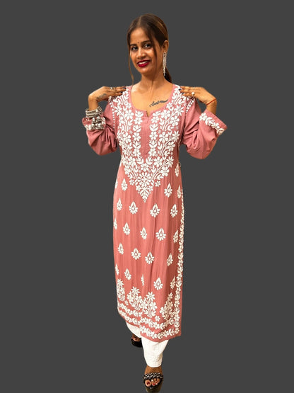 A woman modeling a dusty pink long kurta with intricate white floral embroidery, paired with white pants and black sandals, set against a solid dark gray background.