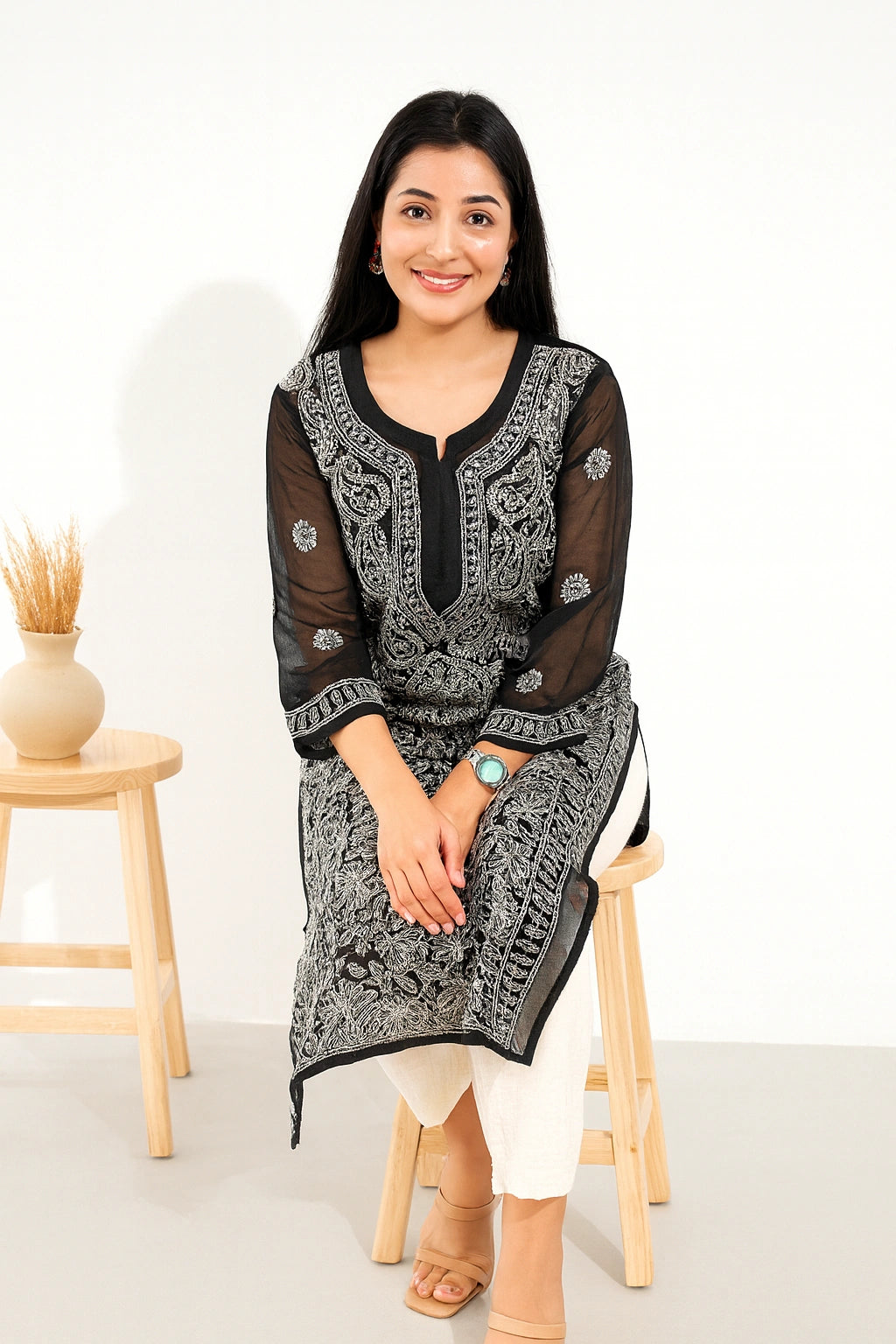 A woman sitting on a wooden stool wearing a black and white embroidered kurta with white pants, set against a clean white studio background with a small wooden side table and vase.