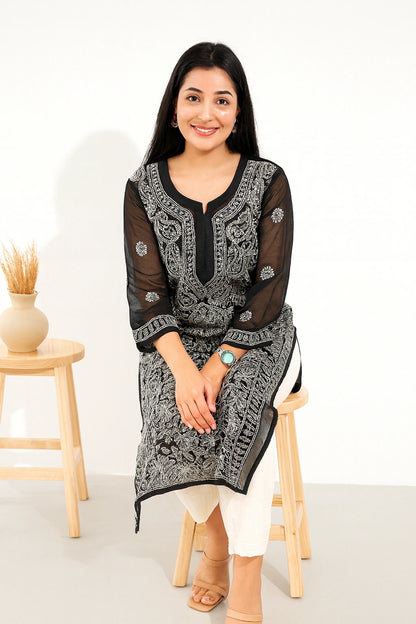 A woman sitting on a wooden stool wearing a black and white embroidered kurta with white pants, set against a clean white studio background with a small wooden side table and vase.