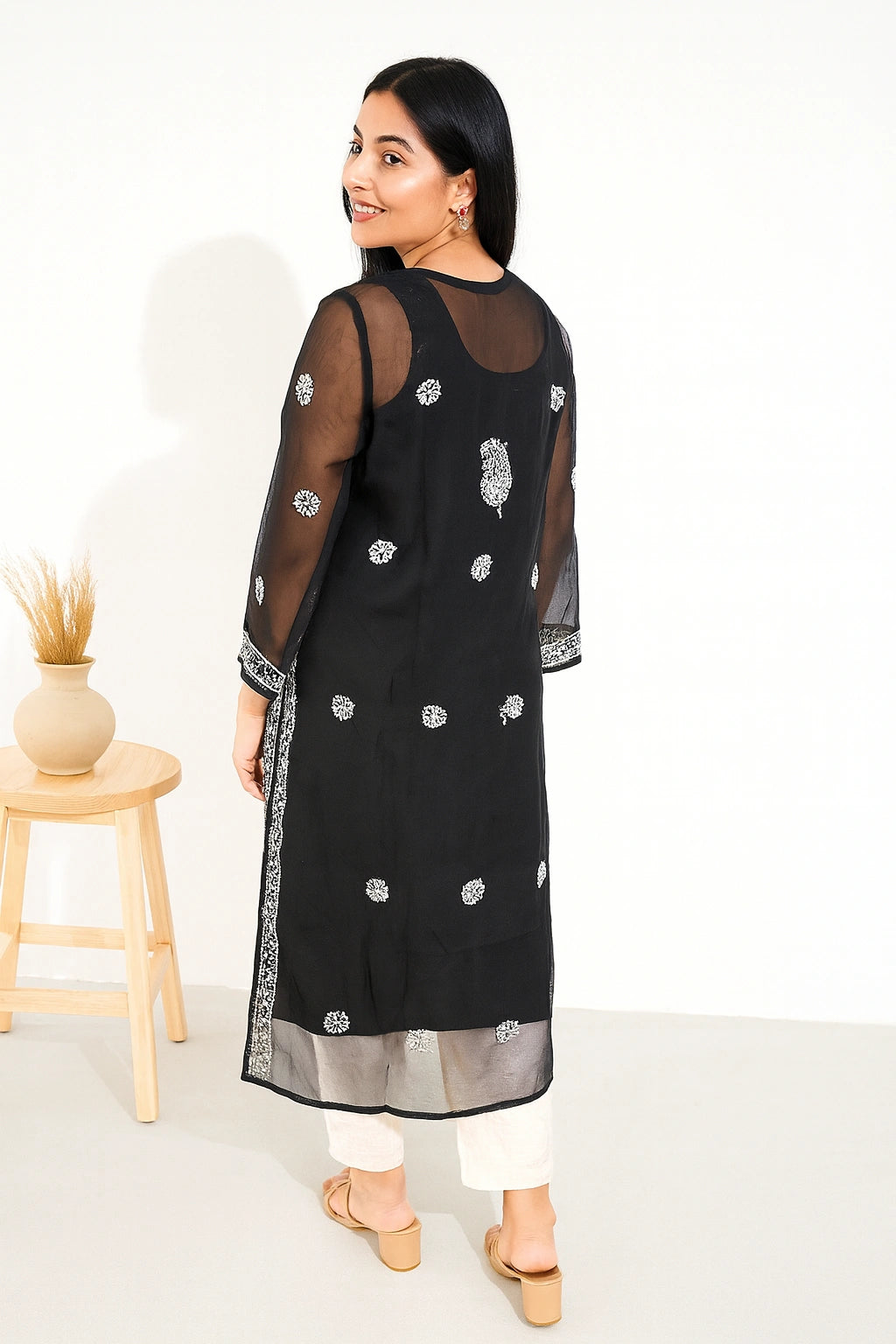A woman modeling a black sheer embroidered kurta with white pants and beige sandals, photographed in a bright studio setting with a small wooden stool and vase at the side.