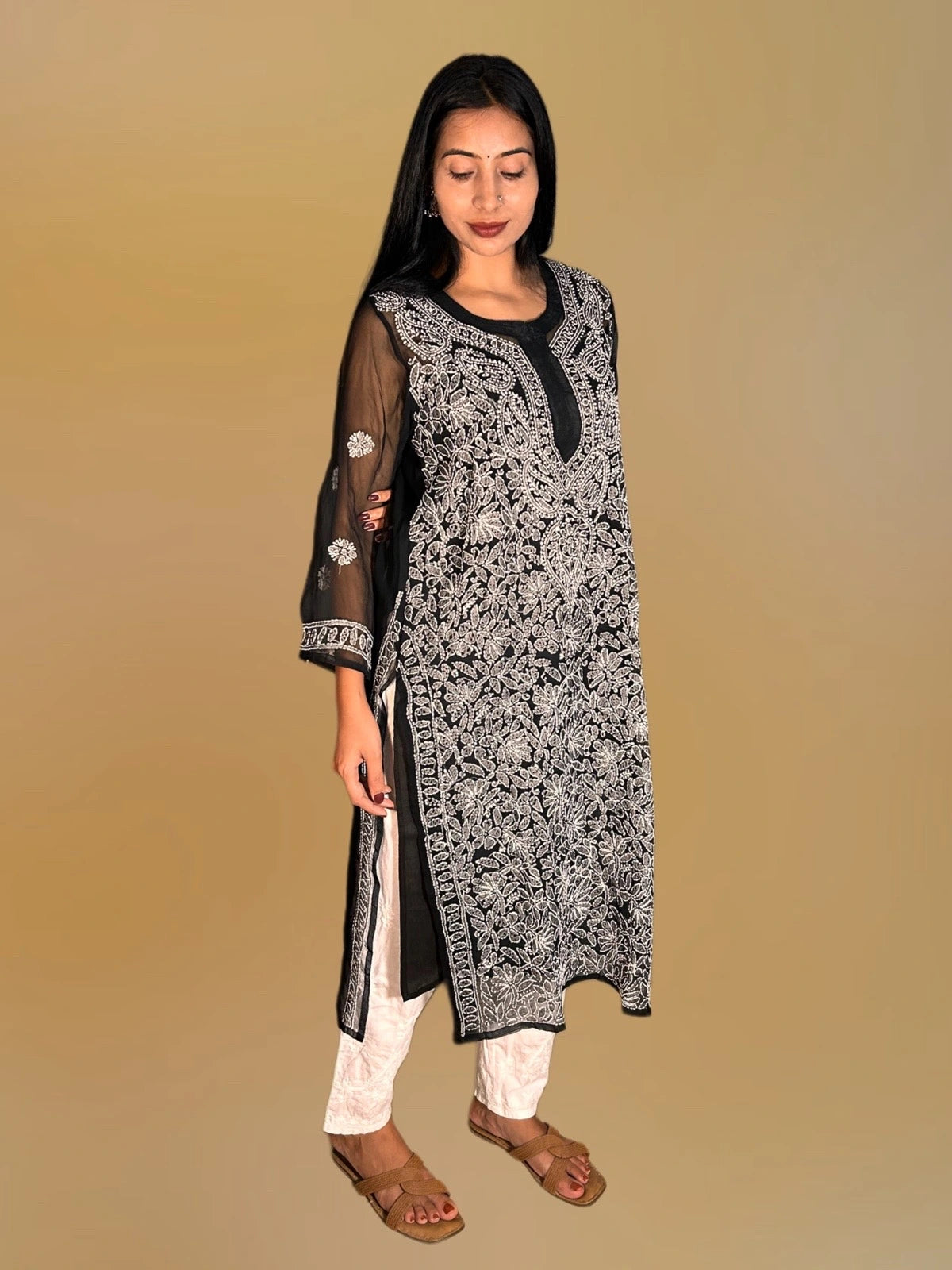Lucknowi chikankari A woman modeling a black and white printed kurta with sheer black sleeves, paired with white pants and brown sandals, set against a solid beige background.