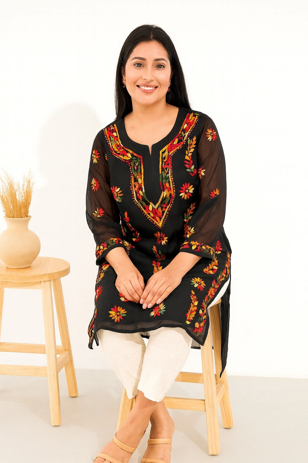 A woman sitting on a wooden stool wearing a black embroidered kurta with red, yellow, and orange floral patterns, paired with white pants and beige sandals.