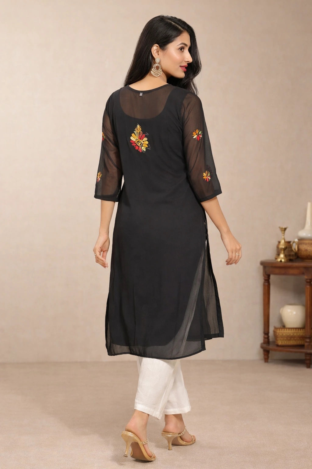 A woman modeling a black sheer mesh kurta with colorful floral embroidery on the back and sleeves, paired with white pants and beige heels in an indoor studio setting.