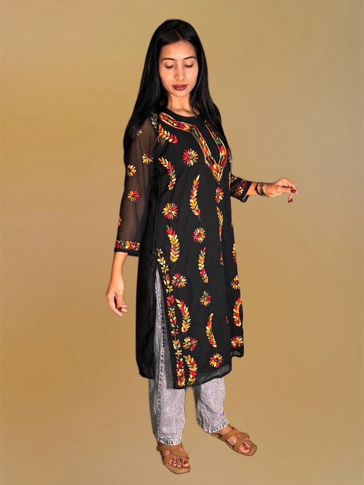 A woman modeling a black sheer long kurta with colorful red, yellow, and orange floral embroidery, paired with light gray jeans and brown sandals, against a solid beige background.