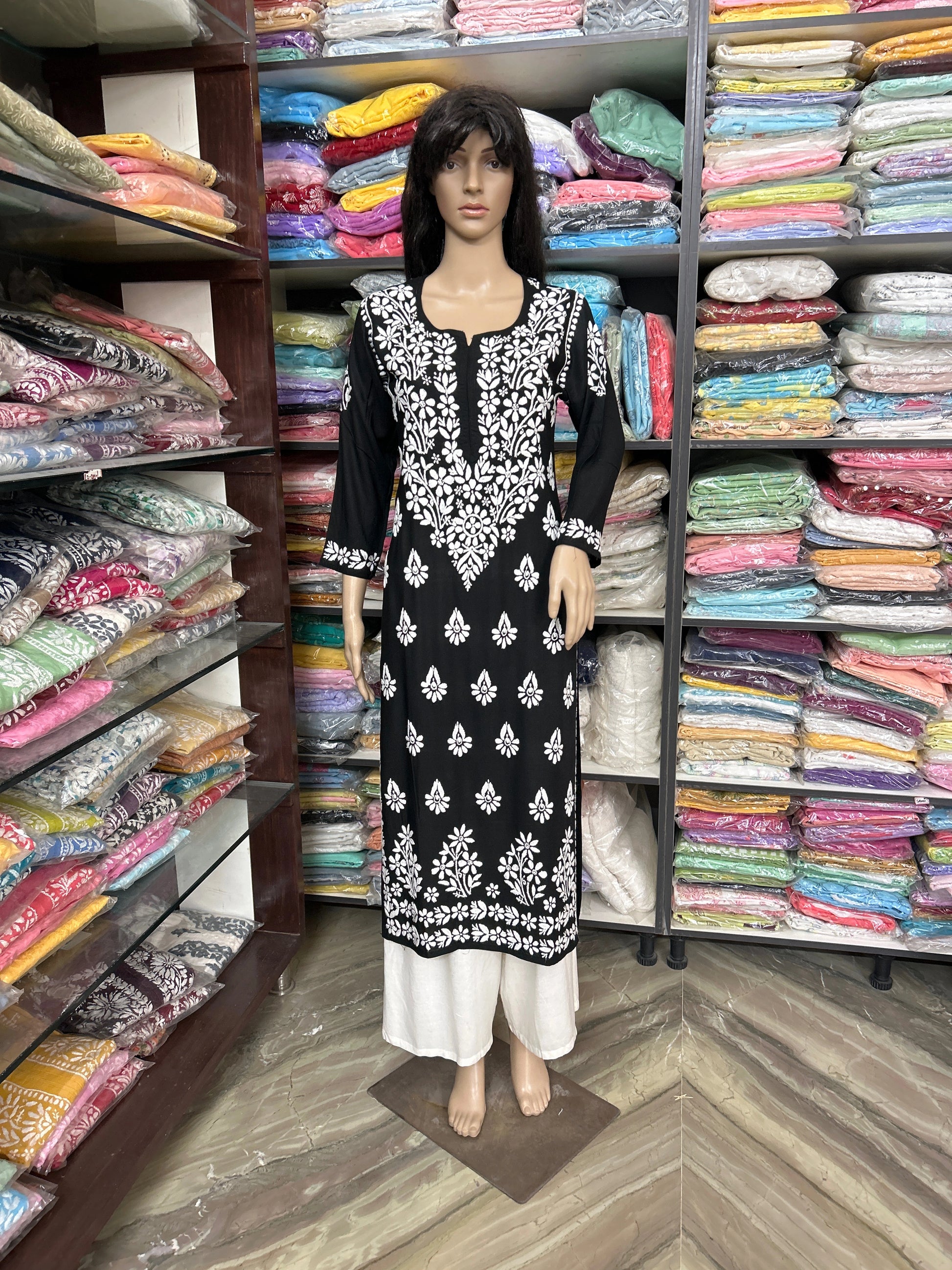 lucknowi chikankari Mannequin wearing a black and white patterned outfit in front of shelves filled with colorful fabrics.
