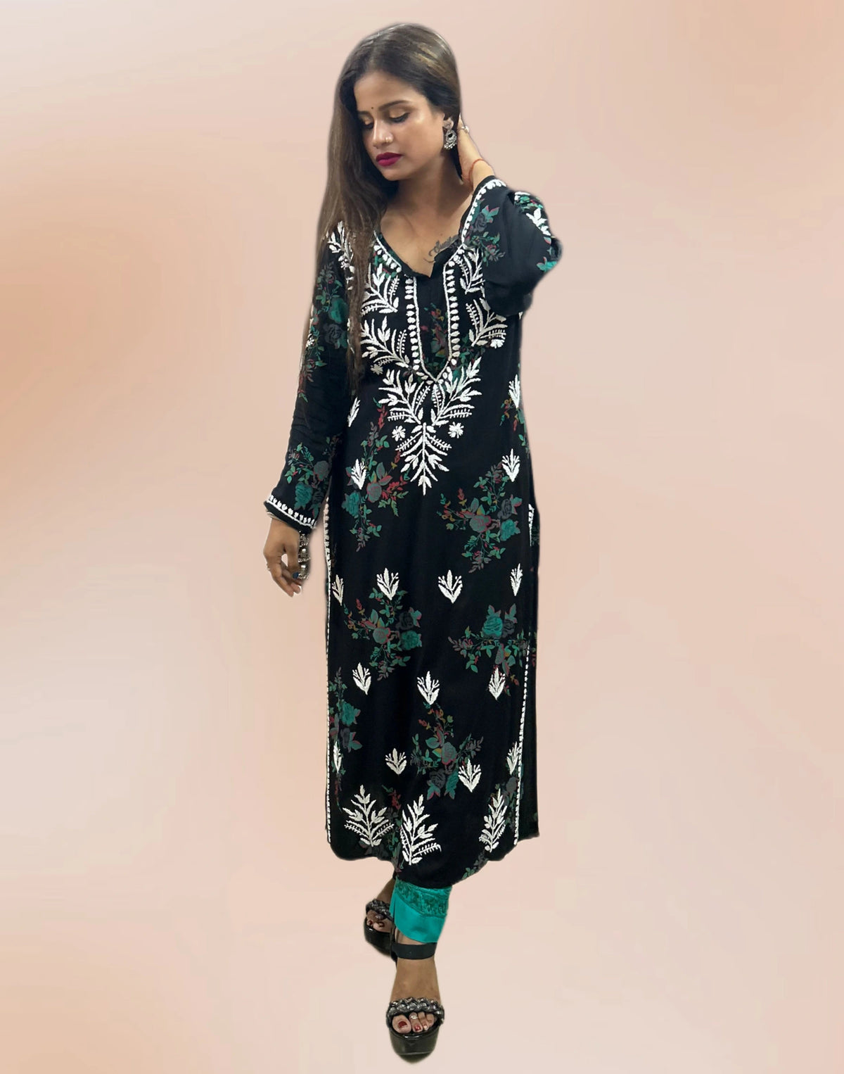 A woman modeling a black long kurta with white and green floral embroidery, paired with teal pants and black sandals, against a solid peach background.
