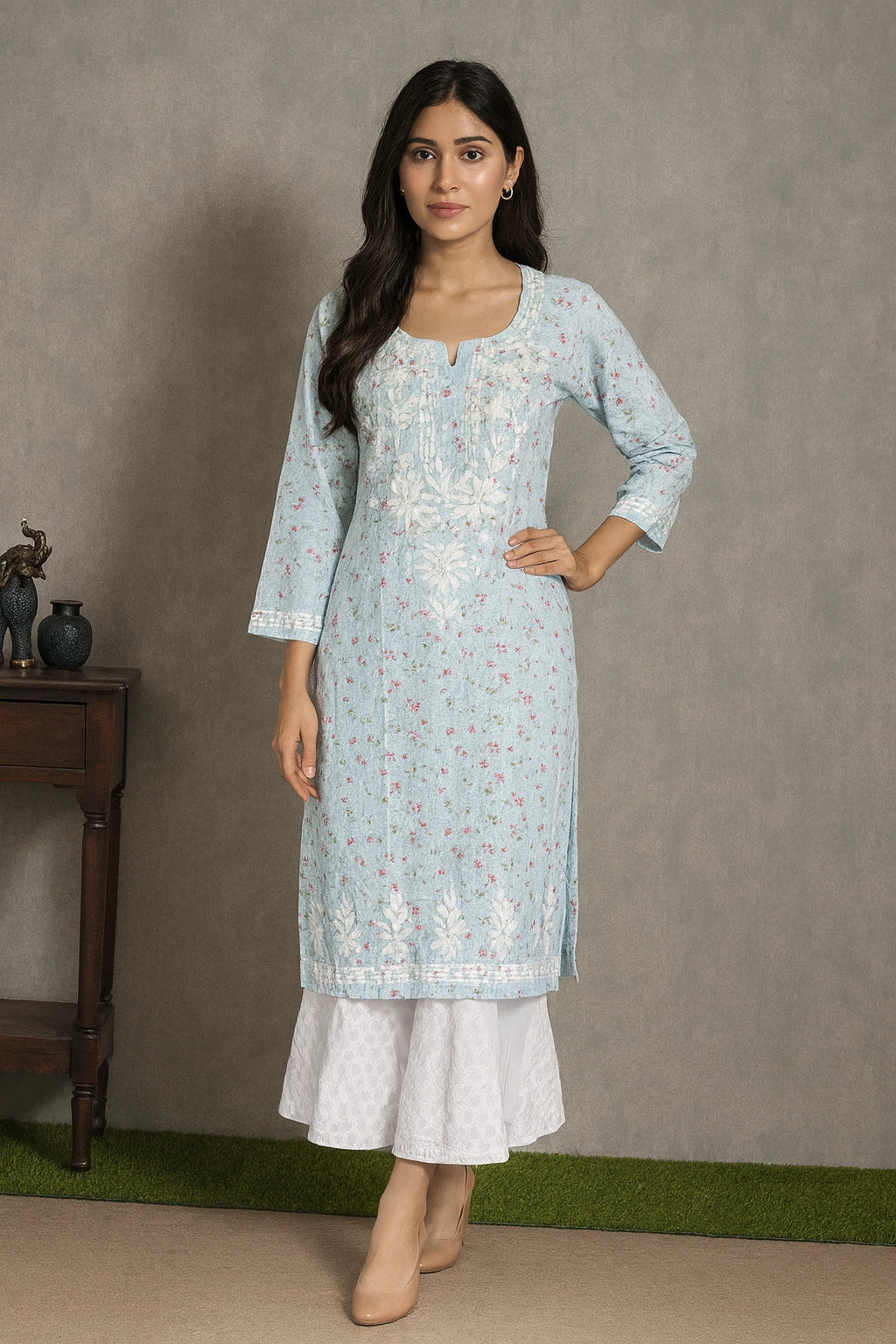 A woman modeling a light blue embroidered kurta with white flared pants, standing against a textured gray wall.