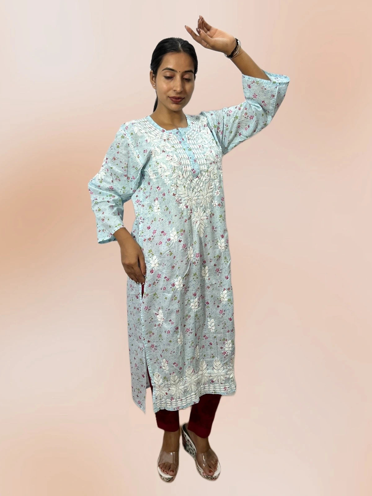 A woman modeling a light blue floral printed kurta with silver mirror work embroidery, paired with maroon pants and clear high-heeled sandals, set against a solid peach-pink studio background.