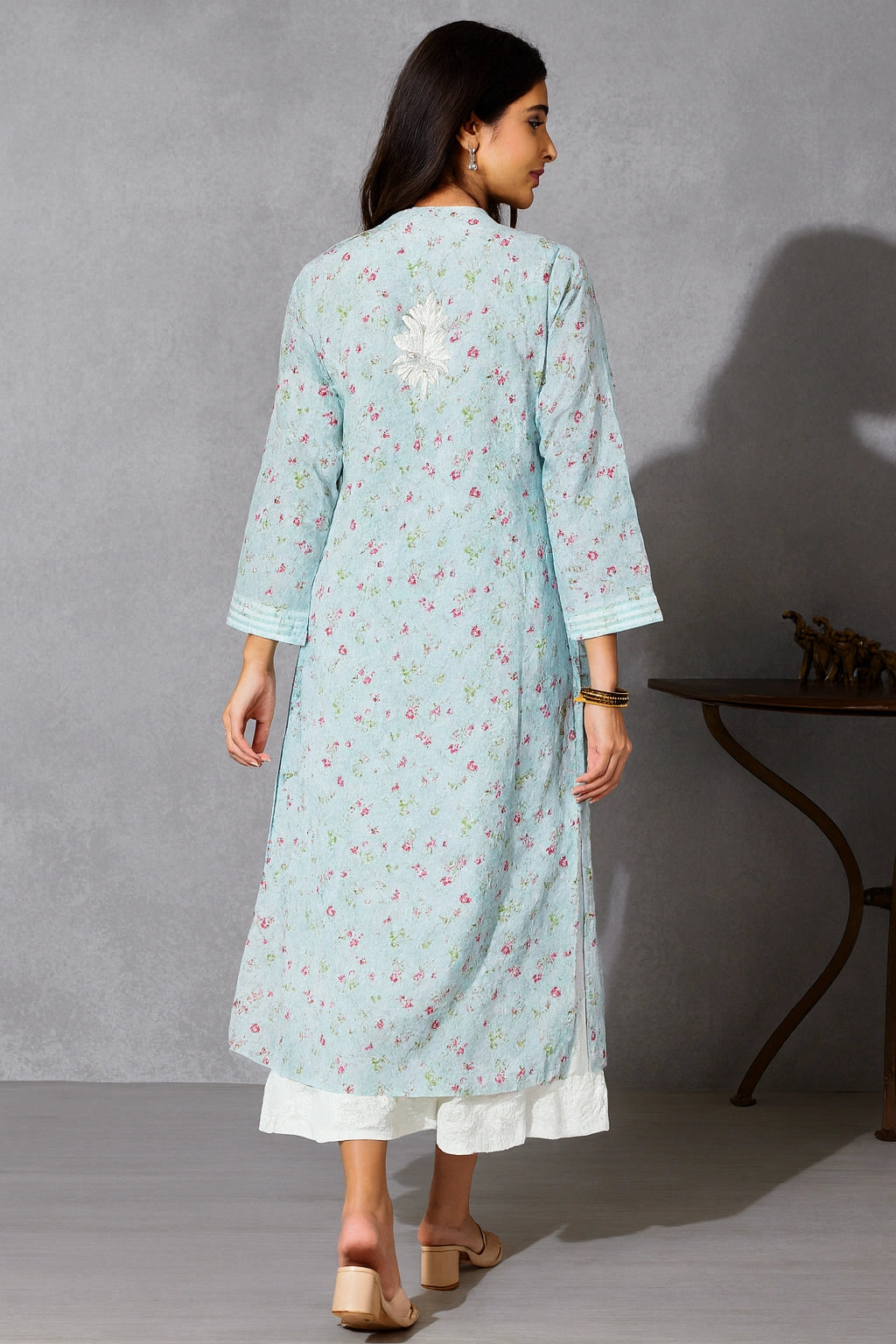 Lucknowi chikankari A woman viewed from the back wearing a light blue floral printed kurta over a white bottom, standing in a studio with a gray textured wall and a dark wooden side table.