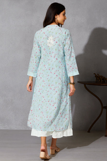 Lucknowi chikankari A woman viewed from the back wearing a light blue floral printed kurta over a white bottom, standing in a studio with a gray textured wall and a dark wooden side table.