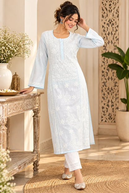 A woman models a light blue long kurta with white floral embroidery and white pants in a warm, decorated indoor setting.