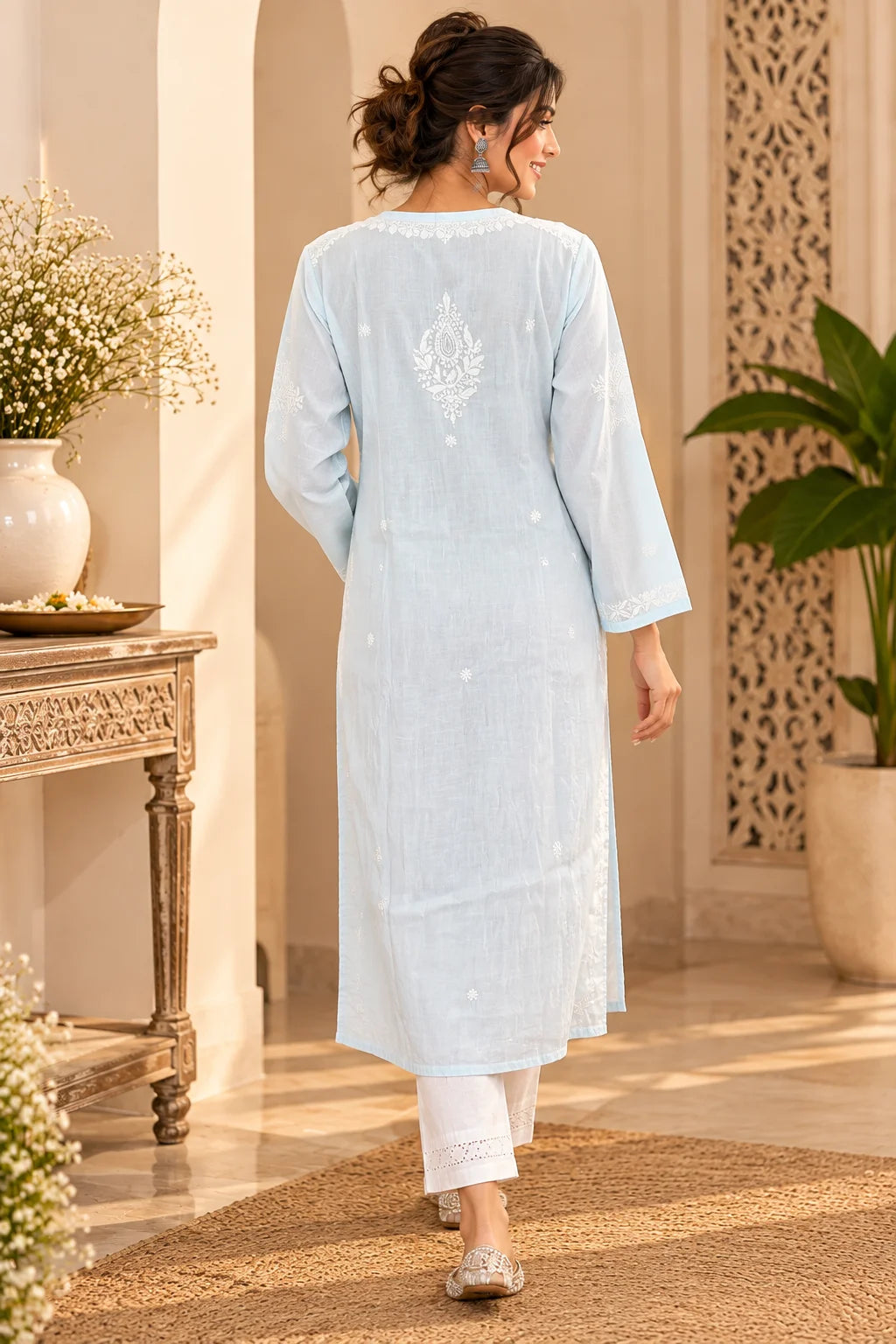 A woman wearing a light blue long kurta with white embroidered details on the back, paired with white pants, walking in a warm, sunlit indoor setting.