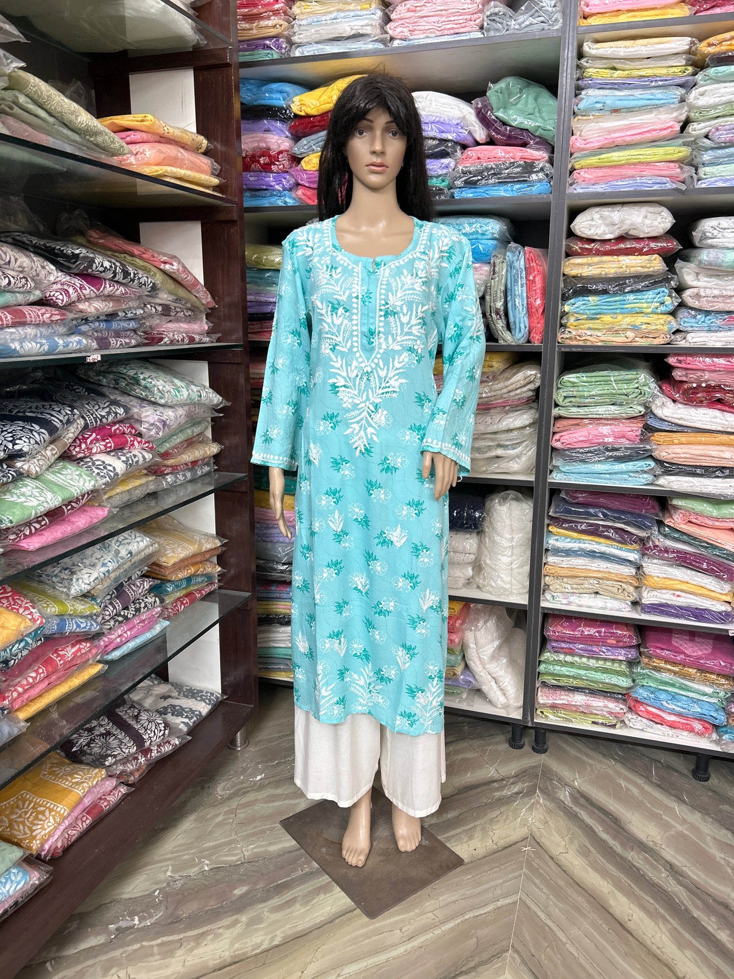 lucknowi chikankari Mannequin wearing a turquoise dress in a fabric store with shelves of colorful fabrics.