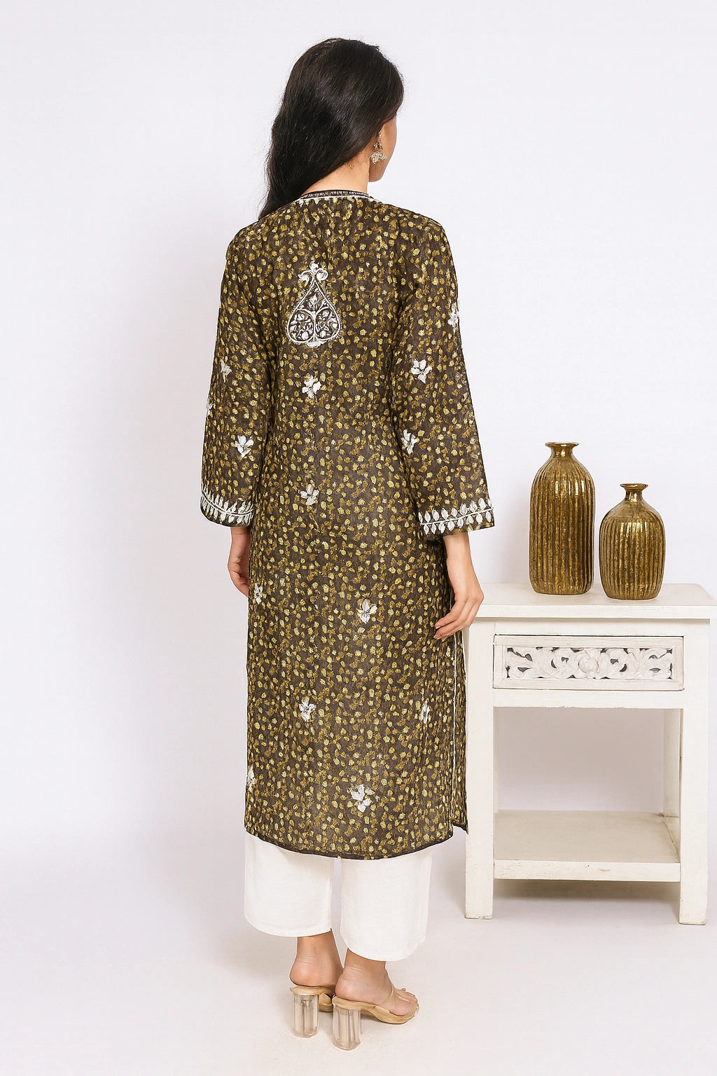 Lucknowi chikankari Woman wearing a green patterned kurta with white pants, standing in front of a white wall with decorative vases.