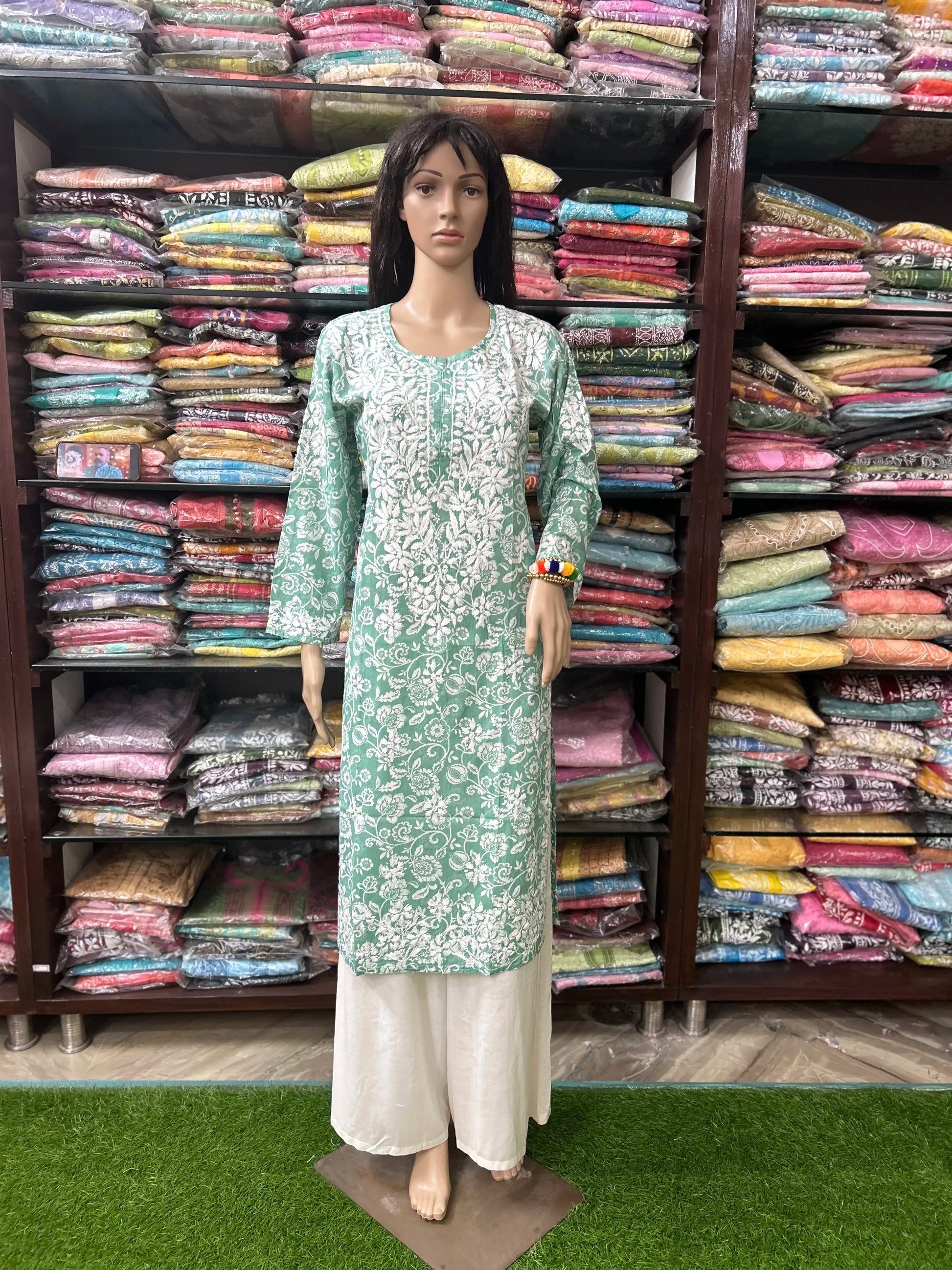 lucknowi chikankari Mannequin wearing a green and white traditional outfit in front of shelves filled with colorful fabrics.