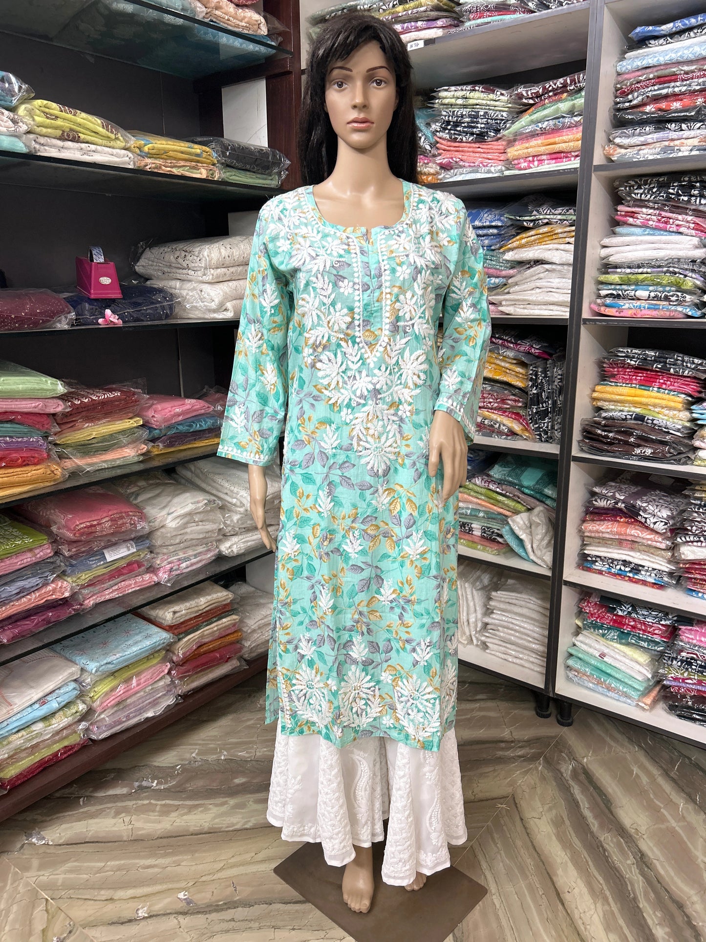 Mannequin wearing a lucknowi chikankari floral dress in a fabric store with shelves of fabric in the background.