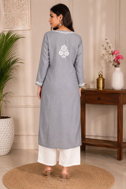 Woman wearing a gray kurta with white embroidery in a room with a wooden table and decorative items.