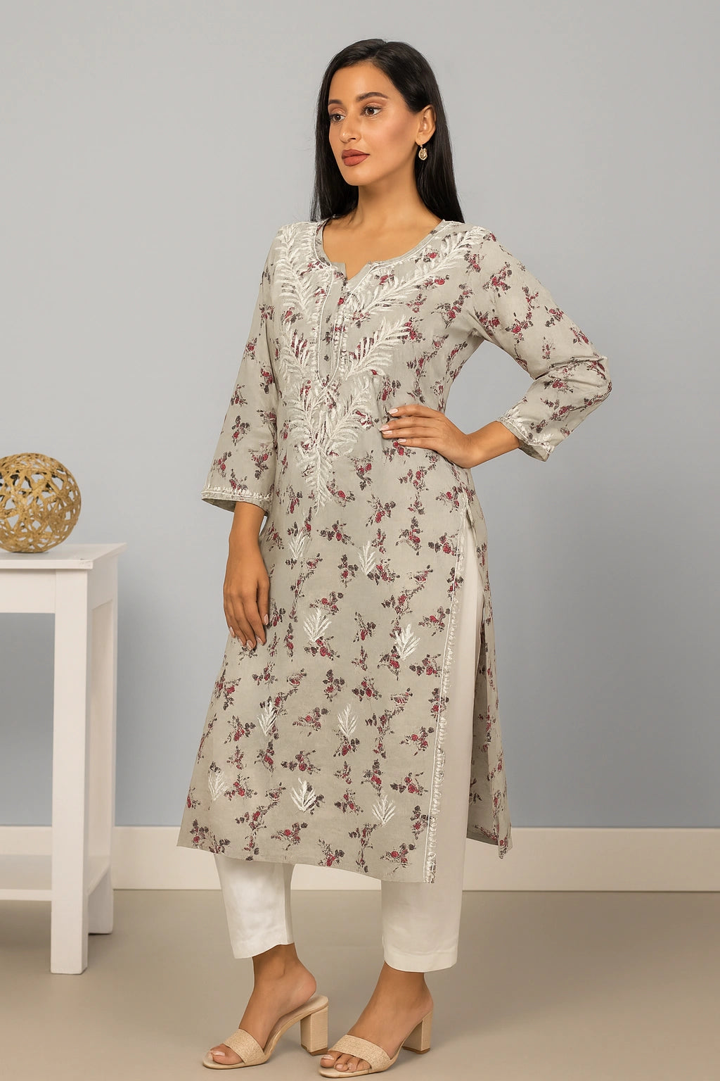 A woman modeling a light beige kurta with white floral embroidery and a subtle brown floral print, paired with white pants and beige heeled sandals, standing in a studio setting.