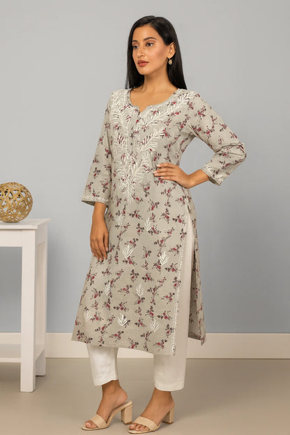 A woman modeling a light beige kurta with white floral embroidery and a subtle brown floral print, paired with white pants and beige heeled sandals, standing in a studio setting.