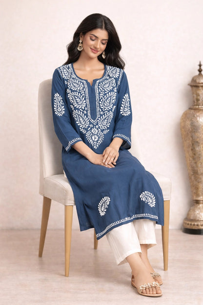 Woman sitting on a chair wearing a blue embroidered kurta with white pants against a beige background
