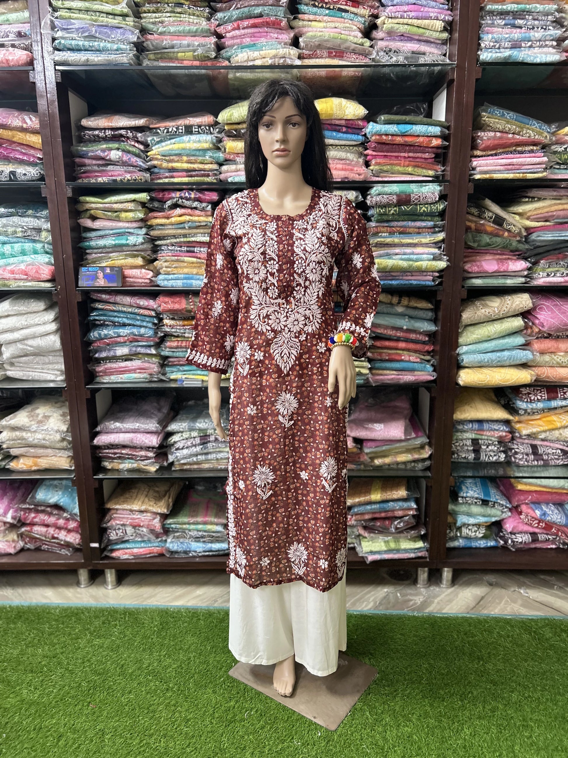 Lucknowi Chikankari Mannequin wearing a patterned dress in front of shelves filled with colorful fabrics.