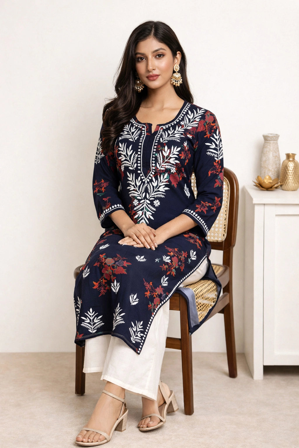 A woman seated on a wooden chair wearing a navy blue embroidered kurta with white and red floral patterns, paired with white pants and beige sandals in a bright indoor setting.