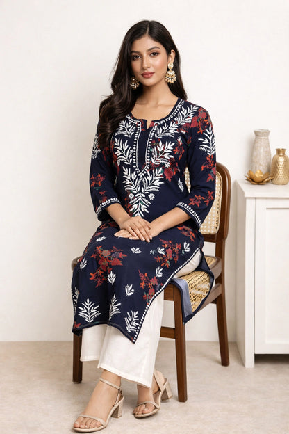 A woman seated on a wooden chair wearing a navy blue embroidered kurta with white and red floral patterns, paired with white pants and beige sandals in a bright indoor setting.