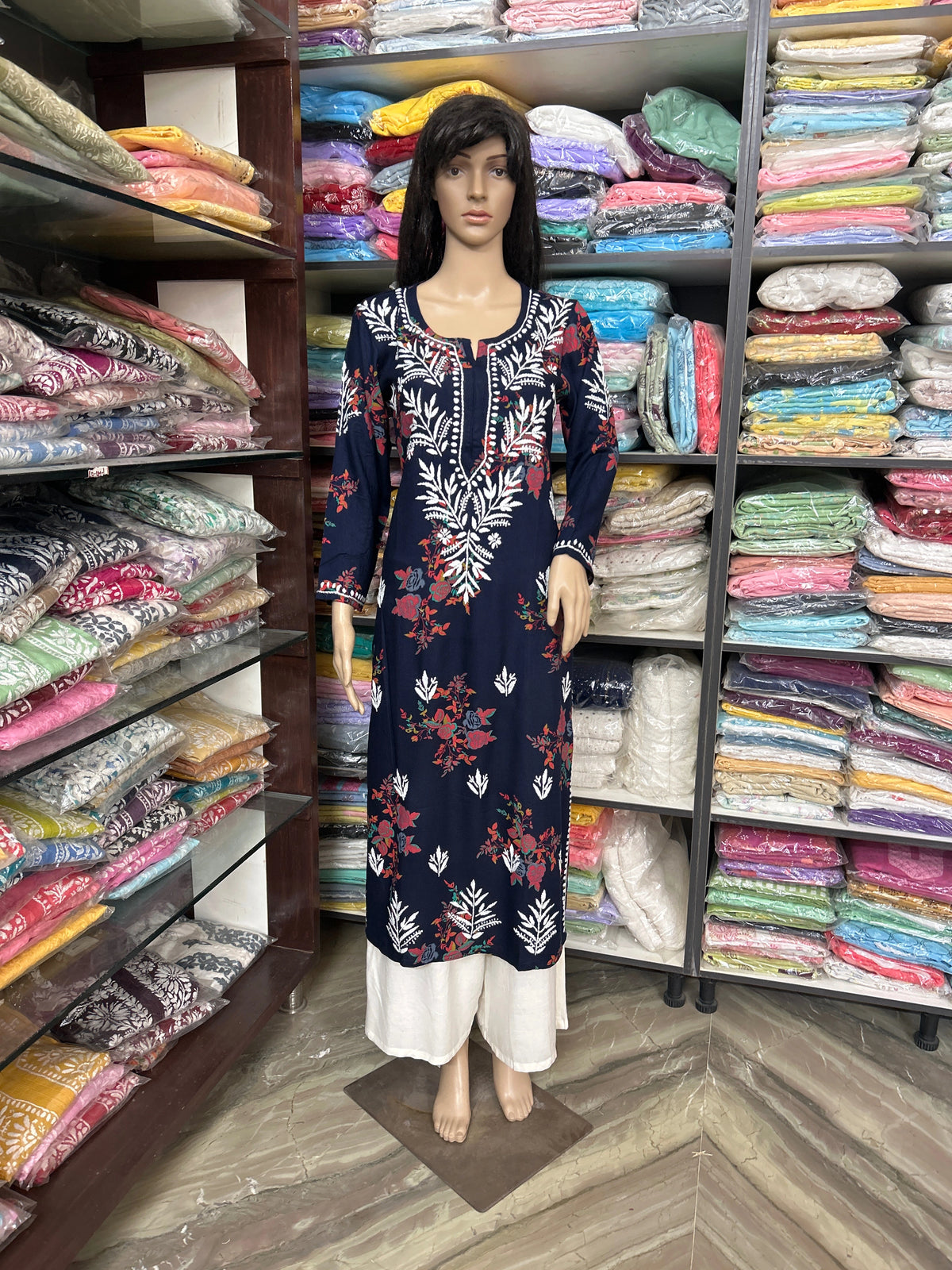 A navy blue long kurta with white and red floral embroidery and print, displayed on a female mannequin in a clothing store. The kurta is paired with white pants, and the mannequin is positioned in front of shelves filled with folded colorful garments.