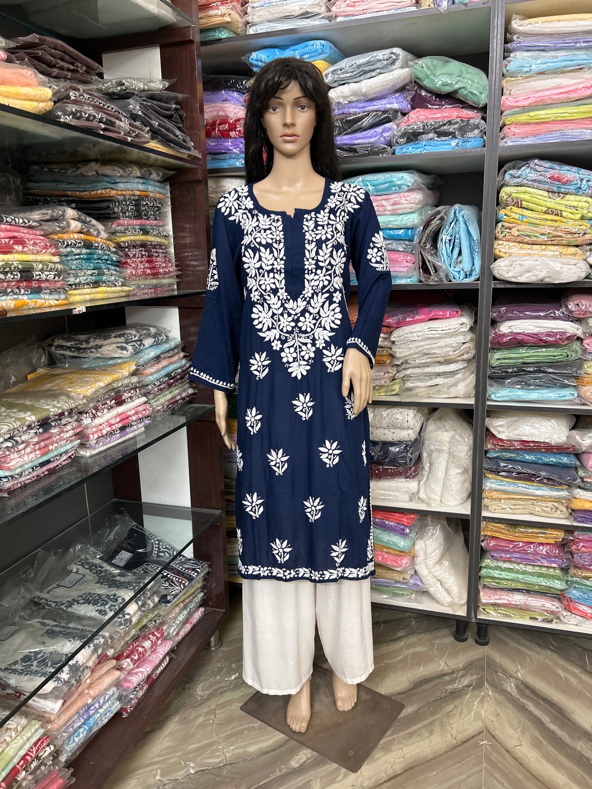 Mannequin wearing a lucknowi chikankari floral dress in a fabric store with shelves of fabric in the background.