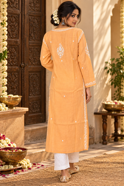 A woman wearing a peach-colored embroidered kurta with white pants, standing in front of a carved wooden door with floral decorations.
