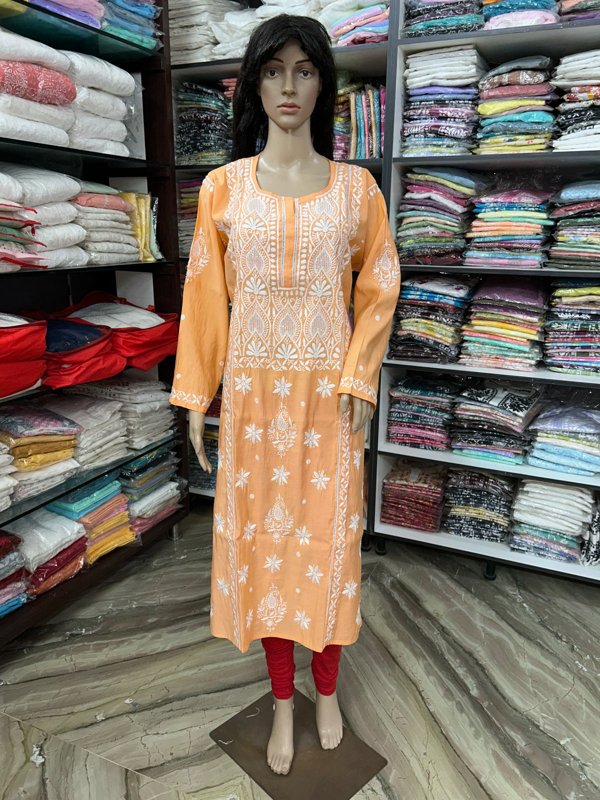 An orange long kurta with white floral embroidery displayed on a mannequin in a clothing store. The kurta features a round neckline, three-quarter sleeves, and white embroidered patterns on the front and cuffs.